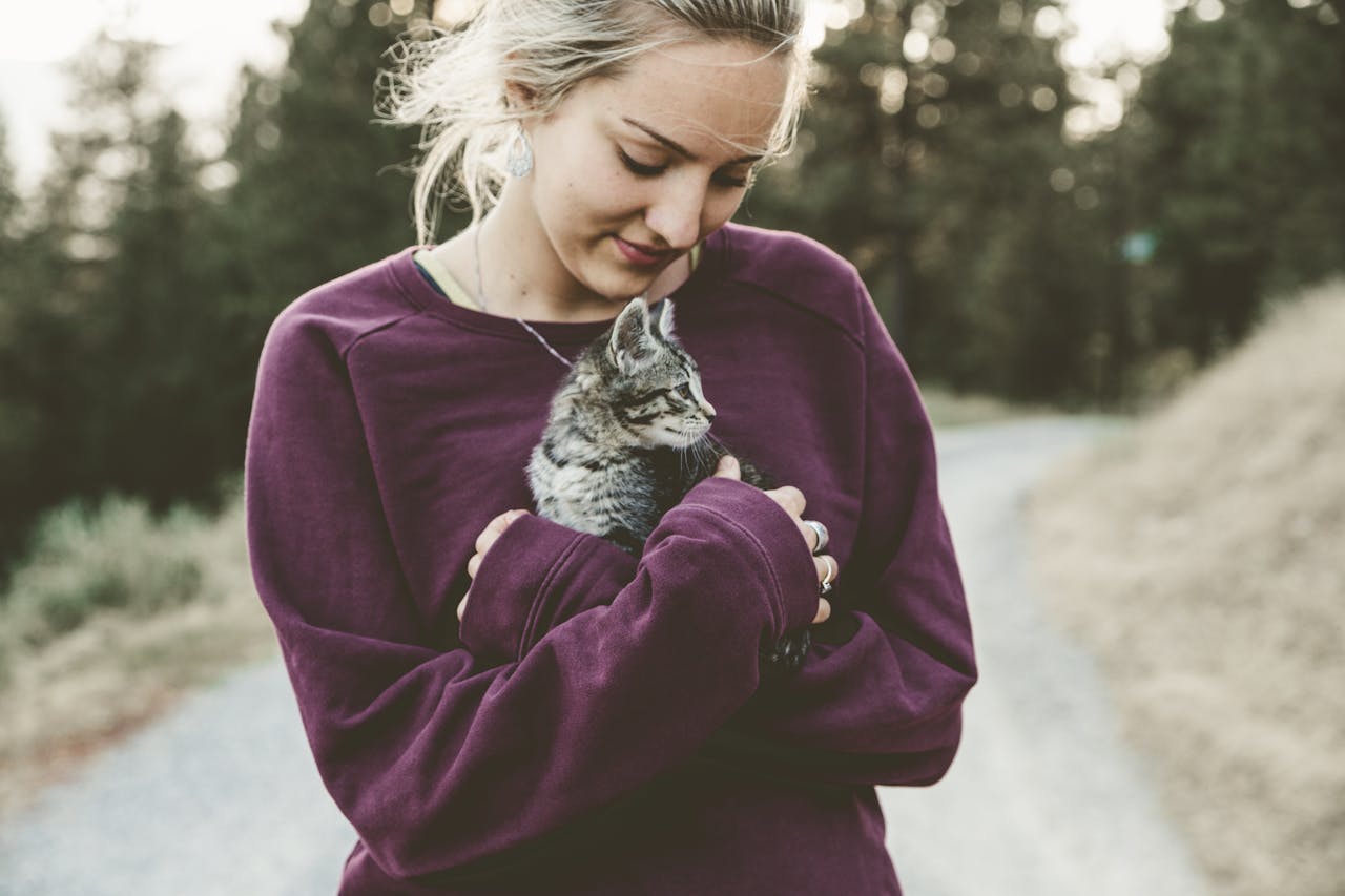 our-services-1 Young woman in a purple sweater smiling down at a kitten she's holding outdoors on a pathway.